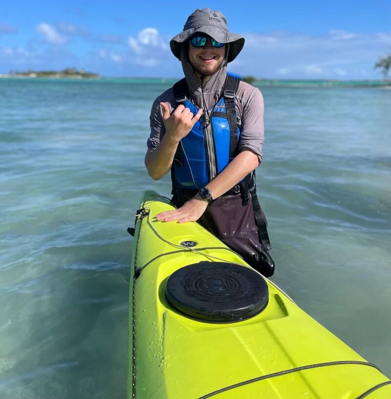 A man wearing a hat, sunglasses, and a life vest is on a yellow kayak in the ocean. He is smiling and giving a shaka sign with his right hand. The water is clear and blue-green, and there is an island in the background. The sky is blue with some clouds. The man appears to be enjoying a sunny day on the water.